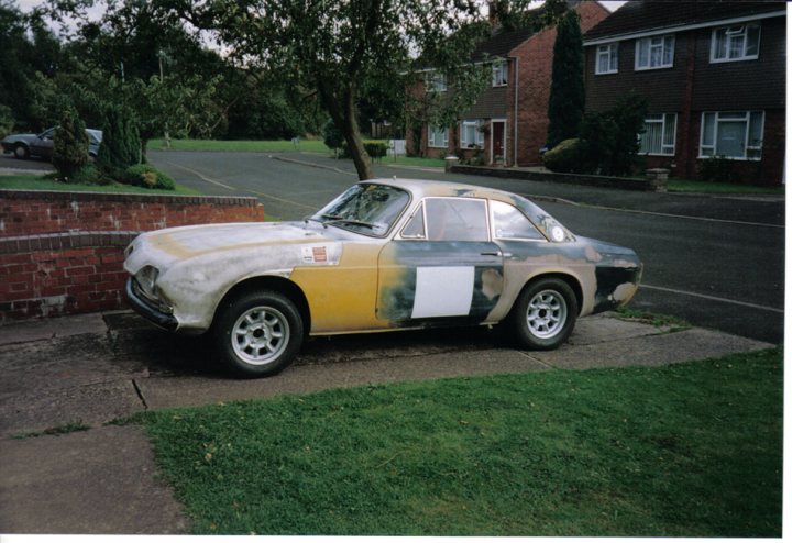 Scimitar Pistonheads Reliant - The image features an old, weathered car parked on a concrete driveway. The car's paint is faded and peeling, indicating age and exposure to the elements. The car is parked in front of a red brick wall and a lush green lawn, giving the scene an urban garden feel. In the background, two-story residential houses can be seen, suggesting that this image was taken in a residential area. The flat lawn and the neatly parked car on it indicate a well-maintained neighborhood.