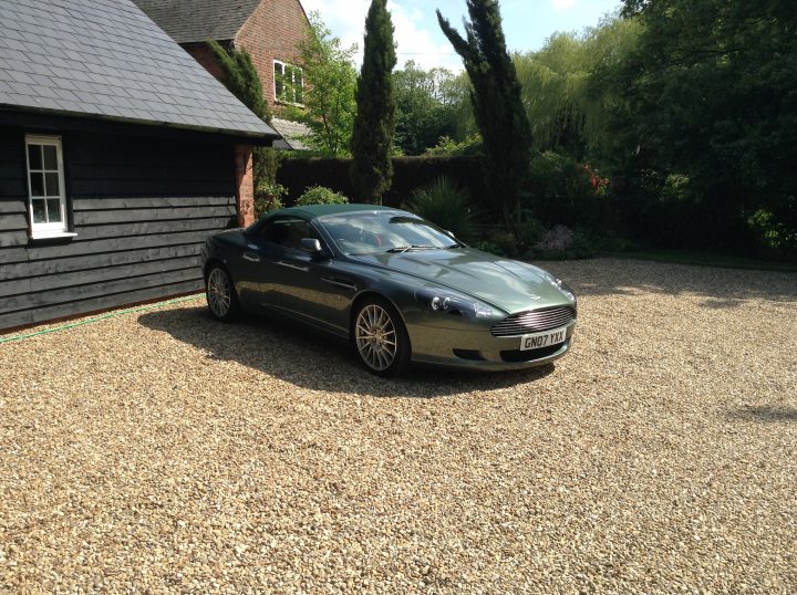 Show us your DB9  - Page 3 - Aston Martin - PistonHeads - This image showcases a sleek silver Aston Martin DB5 convertible parked on its side in front of a black building. The car is positioned on a gravel driveway, with its shiny exterior reflecting the sunlight. The background features a lush, green landscape dotted with trees and bushes, suggesting a serene and picturesque setting. The overall scene conveys a sense of quiet luxury, amplified by the presence of the sports car. The car's position on the gravel indicates it has been recently driven.