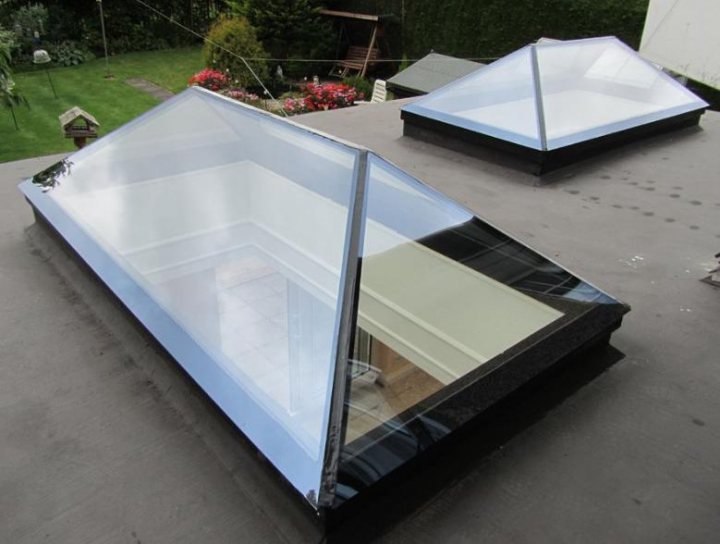 Frameless Skylights Pistonheads - The image displays a unique outdoor living space, likely a patio or deck, featuring two transparent infinity-edge pools embedded into the gray concrete surface. The pools have curved glass designs that appear to extend beyond the physical boundary, blending seamlessly with the surroundings. The area outside the glass appears to be elevated compared to the stone patio area, hinting at two separate levels of outdoor living. The backdrop of the scene is lush greenery, with trees and shrubbery providing a natural contrast to the stark, modern design elements of the glasses. The water inside the pools is clear and still, reflecting the outdoor surroundings and enhancing the surreal, dream-like quality of the setup.