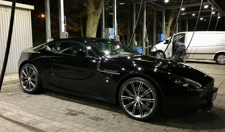 20" DBS wheels on '06 V8 Vantage? - Page 1 - Aston Martin - PistonHeads - The image depicts a black sports car parked at a gas station. The car is sleek and shiny, reflecting the lights from the gas station and the street lights. It's parked next to a diesel pump, which is accessed by a man wearing a black jacket. There are other vehicles and a visible sign behind the car. The backdrop features a roadway with streetlights illuminating the scene.