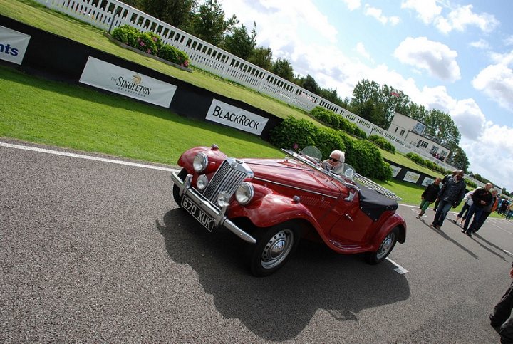 Goodwood Breakfast Club, Pre 1966 classic sunday - Page 1 - Goodwood Events - PistonHeads - The image captures an old-world charm with a red antique car parked on a gray tarmac area with patches of grass. The car's rich maroon color stands out against the softer green and gray background. Two people can be seen in the vicinity of the car, one of whom appears to be its driver, dressed in a cap. Behind the car, a man with white hair is also visible, seemingly maintaining the car's vintage appearance. The setting likely belongs to an indoor auto exhibition, as indicated by the visible banners and the presence of other visitors in the background.