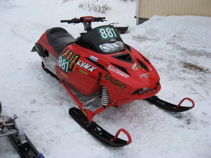 The image depicts a snowmobile parked on a snowy surface. It is bright red and has the word "LYNN" on its side and a number "881". The snowmobile likely belongs to a person named Lynn. The frame of the snowmobile suggests it may be an off-road vehicle, indicating it may be used for recreational snowmobiling or as a vehicle for navigating snowy terrains during the winter season.