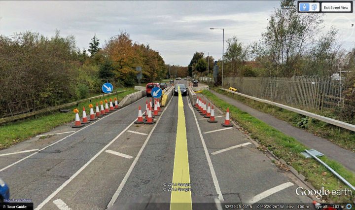 Evil 6'6" width restrictions - Page 9 - General Gassing - PistonHeads - The image depicts an autumnal outdoor scene with a construction area. The focal point is a narrow road with various signs and traffic lights, suggesting work is taking place or adjustments to traffic flow are in progress. Along the sides of the road are multiple orange and white traffic cones, some of which have reflective material on them. The road is lined with green grass and trees with autumn foliage, enhancing the ambiance of the rural or suburban setting. The sky above is gray and overcast, which could lead to rain or mist. The image is likely taken in transit, as indicated by the map and compass icon at the top right, suggesting a device for navigation or location tracking is being used.