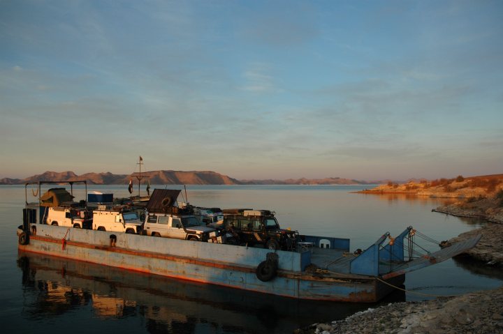 A couple of boats that are sitting in the water - Pistonheads - The image shows a boat resting in still water during what appears to be either sunrise or sunset. The boat is quite large, with multiple layers and a blue top. On the boat, there is a variety of equipment and vehicles that to me seem to belong to some kind of construction or heavy duty work. In the distance, there is another body of water and mountains rising on the horizon. The sky above the viewer is mostly blue with clouds, suggesting an early or late day.