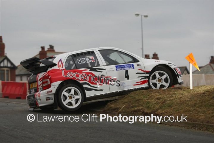 Pistonheads - The image shows a white and red rally car with the number "4" on its side, positioned on a grassy hill at the edge of an asphalt road. The car is visibly on its side, suggesting it has spun out or been crashed. Surrounding vegetation indicates the location is possibly rural or semi-rural. To the right of the car, there is an orange caution flag which is typically used in race events. The style of the vehicle, particularly the roof spoiler and the staging, suggests that this could be a recording set-up for a media shoot, possibly for a motorsport-related event covered by a photography company.