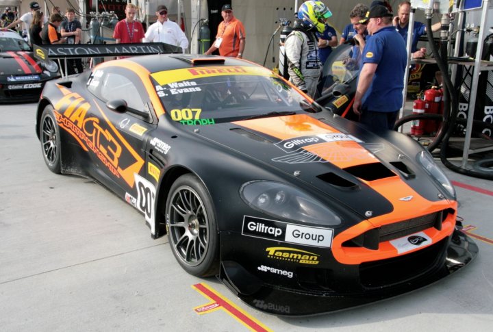 A car that is sitting in the street - Pistonheads - The image shows a race car prominently, predominantly black in color with orange and red accents. The hood is open, and there are sponsorship stickers on its sides, including "Giiltis GB," indicating the brand associated with the car. The license plate number "2007" is visible, suggesting a particular year of racing activity. The car is parked on a track, and there's a group of individuals standing near it, possibly team members or crew discussing the car before an event.