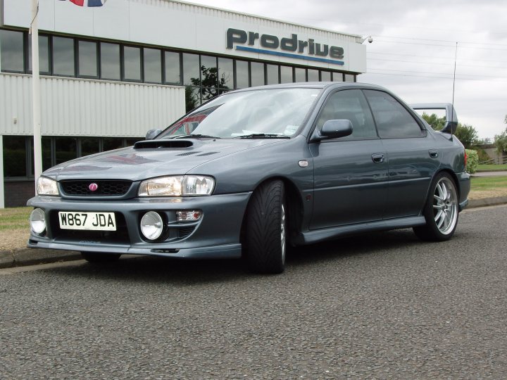 Pistonheads Impreza - The image features a dark-colored two-door coupe parked on a paved surface. It is a sports saloon model, which is visually accentuated with two aftermarket podium lights. In the background, there's a building with the word "PRODRIVE" prominently displayed on the wall, suggesting that the photo might have been taken near a business related to car customization. Additionally, partially visible or out of focus are other cars, indicating that the location might be a parking lot or dealership.