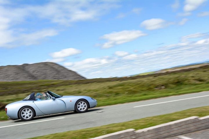 Pictures of your Classic in Action - Page 23 - Classic Cars and Yesterday's Heroes - PistonHeads UK - The image depicts a scene of tranquility and adventure. A silver sports car is driving down a winding, rural road. The car's sleek design suggests speed and performance, while the driver appears to be enjoying the journey. 

The backdrop of this image is a picturesque landscape featuring green hills and a distant mountain range, under a clear blue sky. The road itself curves gently, indicating that it might be a popular route for drivers seeking a thrilling drive amidst natural beauty. Overall, the image conveys a sense of freedom, excitement, and appreciation for the outdoors.