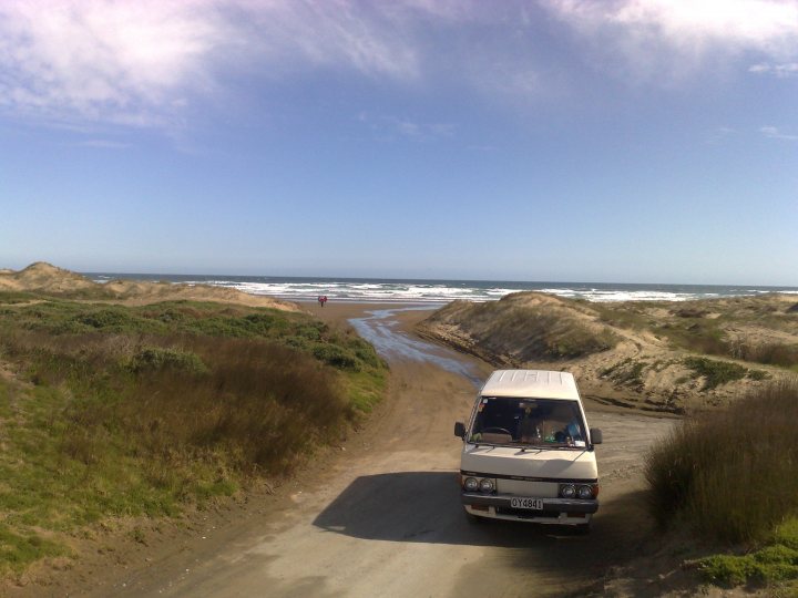 Beach Pistonheads Mile - The image captures a truck driving on a dirt road towards the ocean. The vehicle is located in the lower right quadrant of the image. Above the truck, a coastal road stretches out into the horizon, running parallel with a sandy beach. The sky above is mostly clear but with a few light clouds scattered across it. In the wet grass on the left, there's a hint of greenery. The overall atmosphere gives a sense of adventure and anticipation for the journey towards the beach.