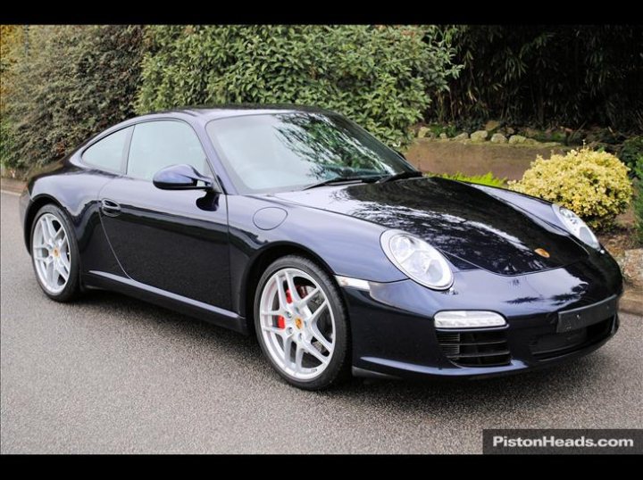 First Test Drive 997 S PDK - Page 1 - 911/Carrera GT - PistonHeads - A sleek black Porsche coupe is the central focus of the image, parked neatly on the street. The car is captured from the side, showing its aerodynamic design and shiny exterior. In the background, situated behind a stone wall and partially obscured by lush green foliage, is a yellow flowering bush. The setting appears to be a suburban or urban area with well-maintained landscaping. The Porsche's positioning suggests a calm, unhurried atmosphere.