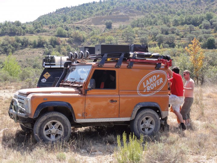 Bought a Orange Defender, Long shot, anyone recognise it?  - Page 1 - Off Road - PistonHeads - In the image, three men are seen inspecting an orange safari vehicle parked in a grassy field. The vehicle appears to be in good condition, equipped with a luggage rack on top, suggesting it's ready for an adventure. Two of the men are actively looking at the vehicle, one of them grabbing the driver's door, while the third one, presumably the driver, is standing behind them, looking over his shoulder. The backdrop of their surroundings consists of a mountainous terrain with a dirt road leading into the distance. The setting and the vehicle's features suggest they might be embarking on a road trip or an outdoor expedition.