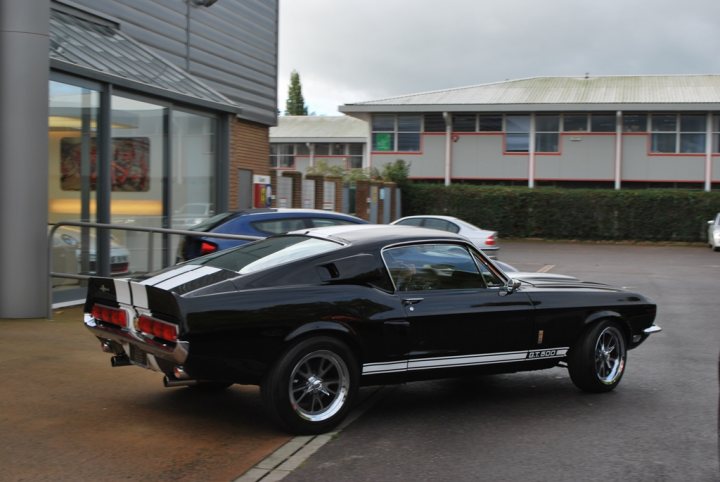 A car parked on the side of the road - Pistonheads - The image depicts a black two-door coupe with a black soft top, parked on an outdoor lot. The car is positioned on the side of a grey concrete roadway next to a building with windows. A few other cars are parked in the background, indicating this might be a shared parking area. There are no visible license plates or distinctive features that stand out from the crowd. The sky is overcast, suggesting a gray or cloudy day.