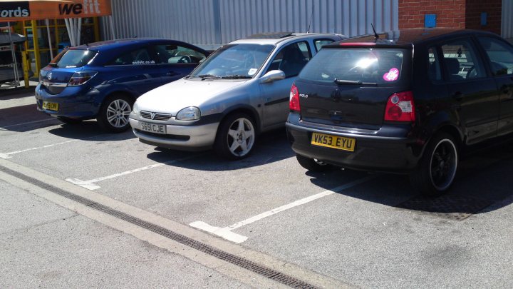 Pistonheads - The image features a parking lot filled with parked cars. There are three cars visible: a black one, a blue one, and a silver one. The black car, a hatchback, is parked towards the right side of the image, while the other two cars are parked a bit to the left. The blue car and the silver one are facing each other in the open space between them.