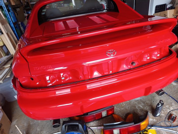 Low Mileage Toyota MR2 MK2. - Page 2 - Readers' Cars - PistonHeads - The image shows a bright red Toyota Corolla car undergoing restoration work. The body of the car is being sprayed with a glossy finish, and it's positioned on a rotisserie stand for a comprehensive paint job. The interior of the car is not visible in the photo. In the background, there appears to be a workshop setting with other cars and equipment, suggesting this is a professional body shop or auto repair garage. There are no visible texts or distinctive markings in the image that provide additional context about the location or the specific model of the car.