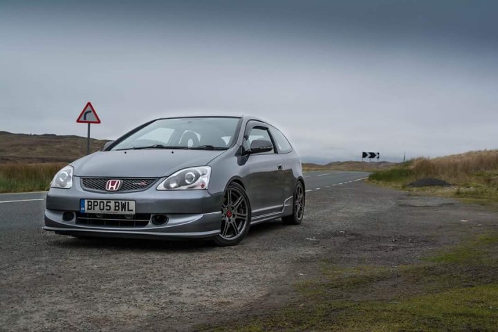 Back in a Honda, EP3 Civic Type R - Page 6 - Readers' Cars - PistonHeads - This image depicts a gray Honda Civic car on a rural road. The car features a distinctive black and red license plate, which reads "BPS BLW." In the background, there is a sign indicating a curve in the road to the left of the car. The setting suggests a tranquil, countryside environment with modest vegetation and muted colors, typical of a rural landscape.