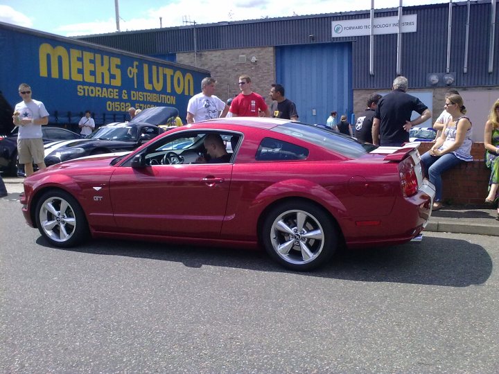 Pistonheads Bbq Sat Atlantic July Sports - This image captures a vibrant street scene with a red sports car as the focal point. The car is driving on the left side of the road, about to enter a parking lot. Several bystanders are standing on the sidewalk, some of whom are having a conversation. The street is lined with various cars, indicating a busy area with a mix of vehicles. The scene is lively, suggesting an urban setting.