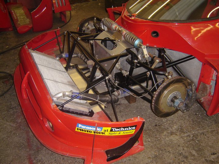 Cooling Pistonheads Problems - The image displays a red button hook vehicle, likely a race car, based on the technical drawings and diagrams attached to the car body. The vehicle is built on a metal frame with various mechanical components protruding. The front end of the vehicle is equipped with a yellow sponge, typically used for absorbing water. The design suggests performance modifications for racing. The text on the vehicle and the stickers visible in the image provide branding and information regarding the modifications. The setting appears to be a workshop or garage where such modifications are carried out.