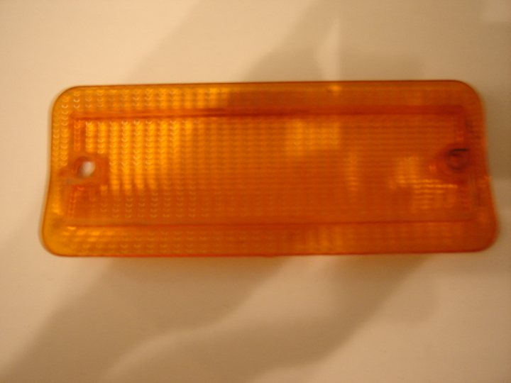 Lenses Indicator Pistonheads - The image shows an orange reflective surface that could be a car mirror cover or a piece of protective clearance on an object. The reflective material is adhered to a white background, and the edges of the reflective surface are not fully aligned with the camera frame, creating a scalloped edge effect. The surface appears textured and is illuminated by a light source from one side, causing some glare and reflections that add depth to the image. The image has a shallow depth of field with the reflective surface in focus while the background is blurred.