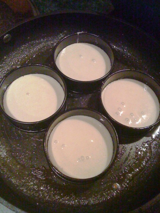 Mums Bikers Hairy Pistonheads - The image depicts a cast iron frying pan with a central burner that's heat-resistant. It's filled with what appears to be a pale-colored dessert or pudding, which could be custard or a similar creamy dish. The pan is sitting on a stove, and the dish looks like it's getting browned, suggesting it might be baking or cooking. There are five distinct desserts or creamy puddings in the pan, each contained in a small saucer. The pan has a matte black finish, which is typical for cast iron cookware, and it looks well-used and seasoned. There's no visible text in the image.