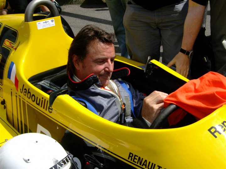 Goodwood FoS pics / videos - Page 1 - Goodwood Events - PistonHeads - The image showcases a man seated inside a vibrant yellow race car. The car is sleek and shiny, its bright yellow body contrasting with the man's gray suit. The man, who is the main focus of the image, is wearing a black helmet and is seen smiling, giving a sense of enjoyment and excitement. His white gloves and the role spotting board with the name "Jabouille" suggest a professional or racing environment. The background, while not the main subject, is filled with additional personnel dressed in black, adding to the overall atmosphere of a professional race setting.