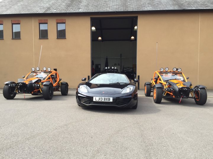 570 / 650 or 12c - Page 2 - McLaren - PistonHeads - This image captures a trio of distinctive vehicles in an outdoor parking area. On the left is a low-lying sports car, displaying a sleek design and black color. Positioned centrally is a monstrous monster truck, marked with the registration number LU3 BX and equipped with large, rounded metal spikes on its sides as a signature element. It sports a vibrant orange and black color scheme. On the right, a third vehicle appears, this one with an unusual tilted to the right, resembling the same monster truck design but in red and black hues. The setting is an open area near a building, characterized by a troupe of hanging lights and a glimpse of a garage or dock.