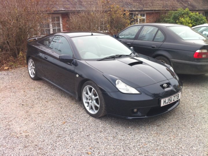 South West PH Car Photos inside... - Page 1 - South West - PistonHeads - The image displays a clean, black two-door coupe parked on a gravel driveway. The text "HJS 1ZT" is visible on the front license plate, indicating the model of the vehicle. Additionally, there's another vehicle with a color contrast partially visible. The setting seems to be a residential area with a house in the background and vacant bushes near the parked cars. The overall condition of the image suggests it might be taken during the day.