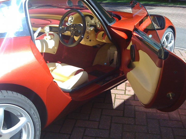 Pistonheads - The image shows the interior of a bright orange or red car, which appears to be a convertible, given the visible soft-top roof. The car's interior is a blend of red and beige colors, with visible seating, the open glove compartment, a standard dashboard, and the instrument cluster that includes a steering wheel and a clock. The car is parked on a brick driveway, suggesting a residential setting or likely a garage or a similar indoor space used for parking. Reflections of light can be seen on the window behind the steering wheel, indicating sunlight shining through the windows, illuminating the car's interior.