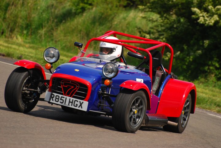 Blue Pistonheads Caterham Kingfisher Viper - In the image, a vibrant two-wheeled vehicle, characterized by its red fender and blue body, is navigating a road lined with trees. The vehicle, equipped with a small windshield and headlamps, appears to be a vintage or unique car design, possibly designed for rally racing given the license plate. The driver, dressed in a white helmet, is engaged in the journey, which is taking place outside following trees that shower a green canopy overhead.