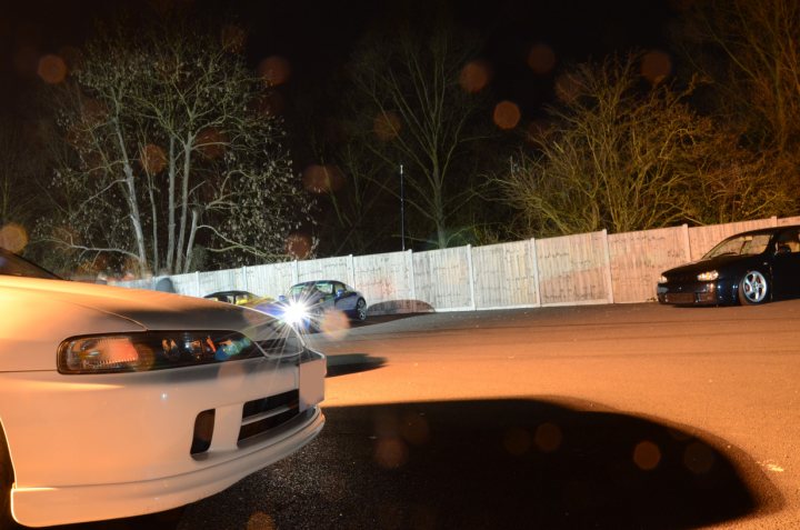 March Essex Pistonheads - The image is a night-time photograph showing two cars parked near each other in a parking lot. The foreground features a white sedan with its headlights on, and the rear of the vehicle reflects the car's lights and the surrounding area in a pattern of spots. The interior light of the other car, a black sedan, is also visible. A white fence can be seen behind the cars, partially obscured by vegetation. The parking lot seems to be empty, giving a quiet ambiance to the scene.