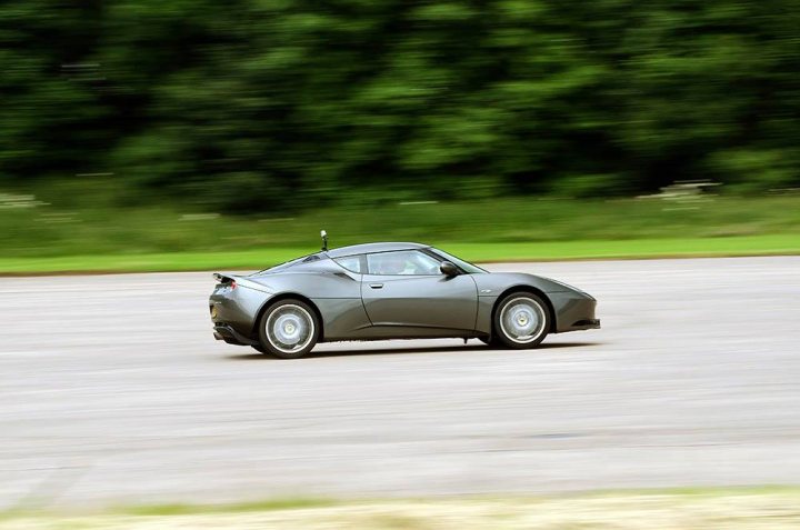 Evora at speed  - Page 1 - Evora - PistonHeads - In the image, we see a sleek, gray sports car, possibly a Lamborghini or a similar ultra-car, speeding down a paved road. The car is captured mid-motion, giving a dynamic sense of speed. The setting is natural, with lush green grass bordering the road and dense trees and foliage in the background, suggesting the car might be moving through a less populated area. The lighting suggests that it could be daytime, but the image has a slightly blurred effect on the background, emphasizing the car's swift movement.