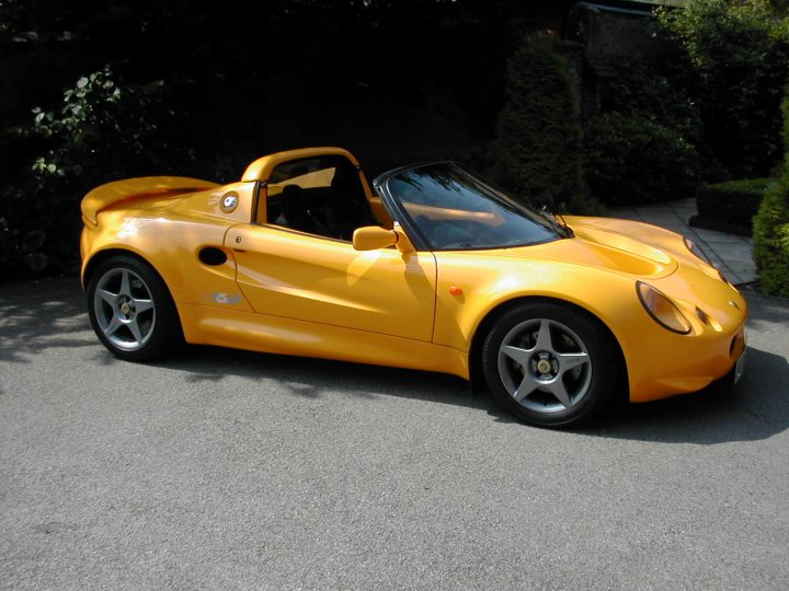 Sport 160 owners - Page 1 - Elise/Exige/Europa/340R - PistonHeads - The image features a vibrant yellow sports car parked on a driveway. The car is a two-seater roadster with a convertible top that is down in this photo. It has a streamlined, curved design with alloy wheels and a prominent front gill. Above the glove box, there's a black entry panel which suggests the car is equipped with a touchscreen system. The surrounding environment includes lush greenery, suggesting a suburban or rural setting. The sky is not visible in the photo, indicating it could be taken during the day or under an overhang.