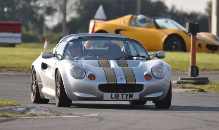 Aesthetics Pistonheads - This image captures a scene of a silver-striped sports car in motion at a race track. The car's aerodynamic body, featuring red accents, suggests high performance. In the background, there's a driver with visible focus, inside a race car cockpit. The background showcases other race cars and the silhouette of fencing, indicating a private racing circuit. Additionally, there's a stop sign visible, reinforcing the exclusively racing atmosphere of the location.