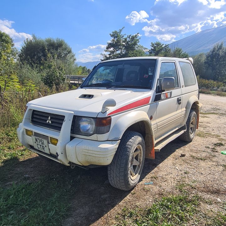 Rare mitsubishi pajero EVO in Albania.  - Page 1 - Japanese Chat - PistonHeads UK - The image shows an old white and red Mitsubishi SUV parked on a dirt road. The vehicle has a noticeable dent on the side, indicating it might have been involved in an accident or hit something hard. It's positioned with a clear sky in the background, suggesting fair weather conditions at the time of the photograph.