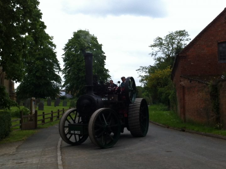 A black and white photo of a steam engine - Pistonheads - In the image, a man in a red shirt is steering a vintage black steam locomotive down a narrow country road. The locomotive, with its large wheels and smokestack, is the central focus of the image. The area is lined with trees and a fence, and there are a few people scattered around, watching the locomotive. The image has a rustic, timeless quality, reminiscent of bygone eras. The scene captures the charm and nostalgia of early steam-powered transportation.