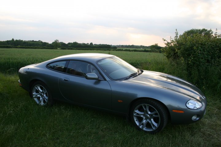 A classic car is parked in a field - Pistonheads - The image features a two-door sports car parked on a grassy area. The car is gleaming and appears to be in pristine condition. It has a steering wheel on the driver's side and is emitting a soft glow from its headlights, suggesting either dawn or dusk. The background consists of rolling hills dotted with trees, creating a serene and picturesque setting. It is a sunny day, as indicated by the soft lighting and the car's reflection on its shiny hood and body.