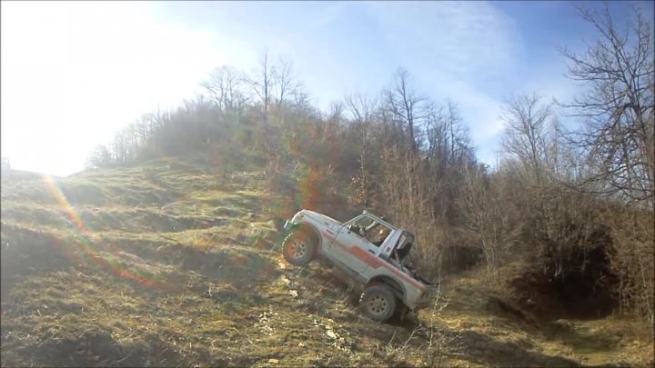 Bit of off-roading, and a bit of camping. - Page 1 - Off Road - PistonHeads - The image shows a white truck balancing on the edge of a steep precipice with a grassy side. The truck appears to be in a precarious position due to the angle it's on. The environment surrounding the truck is rugged and wooded, with a backdrop of clear sky with some scattered clouds. The time of day seems to be midday based on the brightness of the sky. The truck is outdoors and appears to be involved in an off-road driving activity.