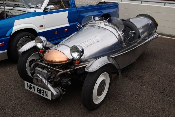 Pistonheads - The image features a sleek, silver and black vintage car situated in a parking lot, adjacent to a bright blue pickup truck. The vintage car is uniquely shaped, with two lights positioned at the front. The vehicle is equipped with wire wheels, a copper-like light diffuser, and a spinning unit. The overall scene is an outdoor setting, possibly a garage or a public parking area.