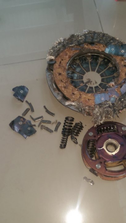 A clock that is on a table in a room - Pistonheads - The image shows a close-up view of an object that appears to be a car spring, which is typically used in the suspension system of a vehicle. The spring itself is caught in shards, indicating it might be broken or fragmented. Scattered around the central spring are small metal bits, possibly pieces of the broken spring. Various other metal components, possibly springs or rings, are spread across the frame, suggesting some disassembly or destruction process is underway or has recently occurred. The setting is a rather nondescript, lightly reflective surface that could be a table or floor.