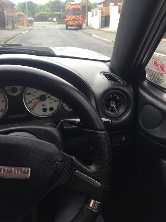 my mx5 - Page 1 - Mazda MX5/Eunos/Miata - PistonHeads - The image provides a close-up view from inside a car looking out at the street. The dashboard of the car is visible, complete with an instrument cluster that includes a speedometer and a cluster gauge performance meter, also known as a racing tachometer, suggesting the car may have a racing pedigree. Outside the car, a road can be seen where a blue and yellow van appears to be pulling up alongside the vehicle. The cabin of the car is made of leather, and there are warning stickers on the vehicle, likely for safety or insurance reasons. The overall scene suggests a small car on a residential street, possibly during a race or high-performance driving situation.