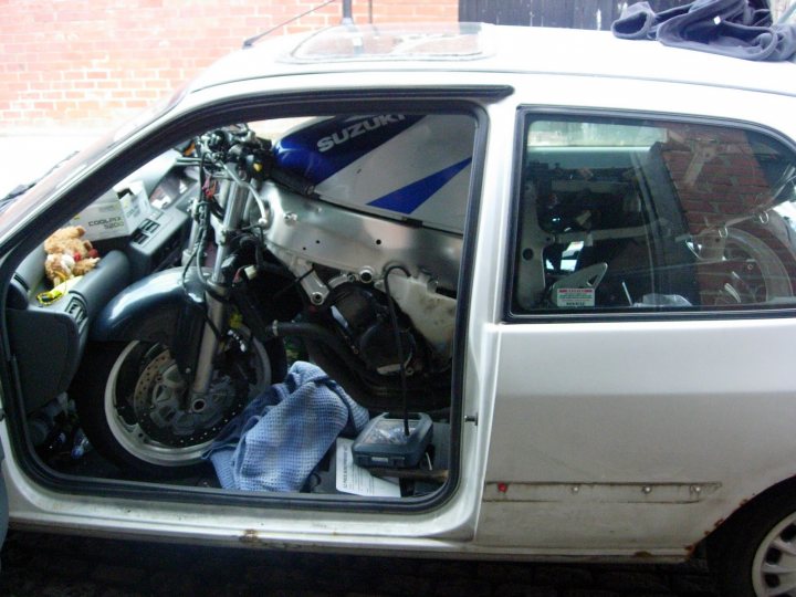 Pistonheads Motorbikbike Boot Fit - The image shows a compact car, parked adjacent to a red brick wall. The car's interior is visible, revealing items like a teddy bear and various assorted items scattered around, possibly belonging to the car's owner. Distinctively, a motorcycle is crammed into the back of the car, occupying the entire trunk area. The motorcycle has a prominent Suzuki logo. A towel can also be seen under the car's backseat, and a few other small items are also discernible within the car. The overall scene suggests a road trip or an active lifestyle where space is used efficiently.