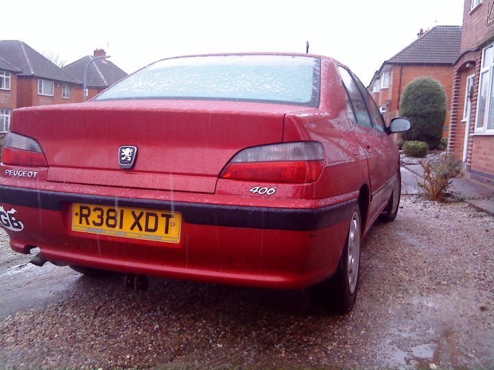 Daily Snotter Pistonheads Free Peugeot - The image shows a red sedan car parked on a residential street. The car is sleek and shiny, indicating that it has been well-maintained or recently polished. The car is branded with the emblem of a lion, suggesting it is a Peugeot vehicle. It has a yellow license plate. The setting appears to be a suburban area during the daytime, with the Glegg in the background, suggesting it might be in a small town or rural area. The street is lined with houses, and there is a visible bush between the cars and the houses.