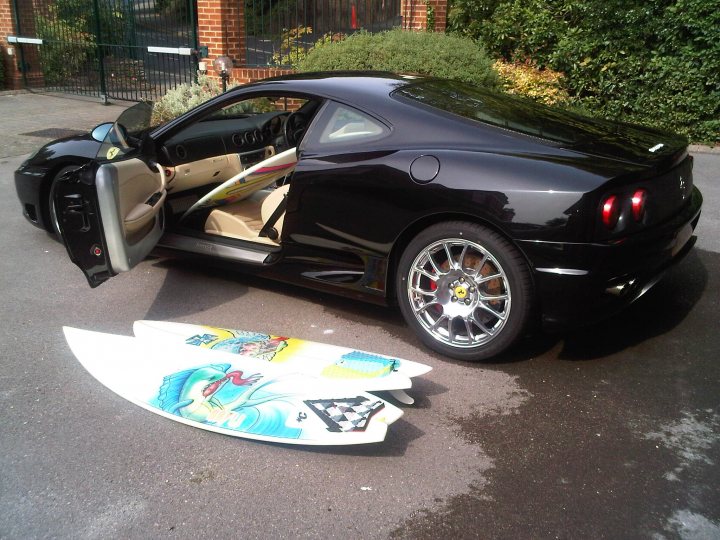 Pistonheads Surfing California Style - The image shows a sleek, black convertible parked on an asphalt surface. The top of the car is currently down, revealing the interior. Lying on the ground next to the car is a surfboard with vibrant graphics. The surfboard is erect, with the upper side facing upwards. In the background, there are green shrubs, suggesting that the car might be parked in a residential area or a parking lot adjacent to a garden or park. The overall scene suggests a day of surfing followed by a relaxing evening or weekend.