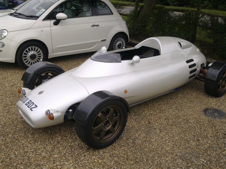 Pistonheads - The image features a vintage-style racing car parked on a gravel driveway. The car is white with black detailing, including the wheel rims. There is another similar but not identical car parked slightly behind it. There are no visible people in the scene, and the setting appears peaceful and serene. The overall impression is of a classic racing car on display.