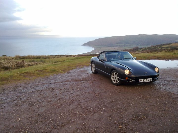 Hi, all you South West folk. - Page 1 - South West - PistonHeads - The image is a photograph depicting a classic black convertible sports car parked on a muddy, uneven surface which suggests it might be near a seashore. The car is shot from a slightly elevated angle, focusing on its interior and the open hood. Behind the car, the outlines of a hilly landscape with sloping fields are visible under a partly cloudy sky. The overall mood of the image is serene and outdoor, blurring the background slightly to keep the car in sharp focus.