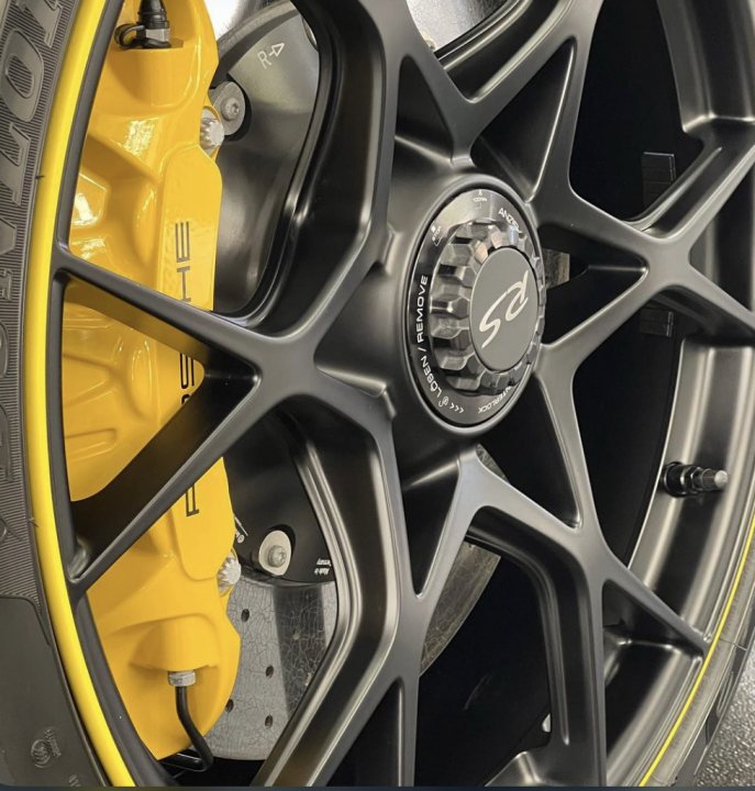 Pistonheads - The image features a close-up view of a car's wheel and its components. The hubcap is black with yellow accents, and the disc brakes are visible behind it. There is a yellow caliper attached to the disc brakes, which contrasts with the other parts in the picture. The wheel has a large bolt that holds the hubcap in place, and there's also a small label or logo on the hubcap. The background of the image is not detailed, but it appears to be a neutral grey color, likely a part of a workshop or garage setting.