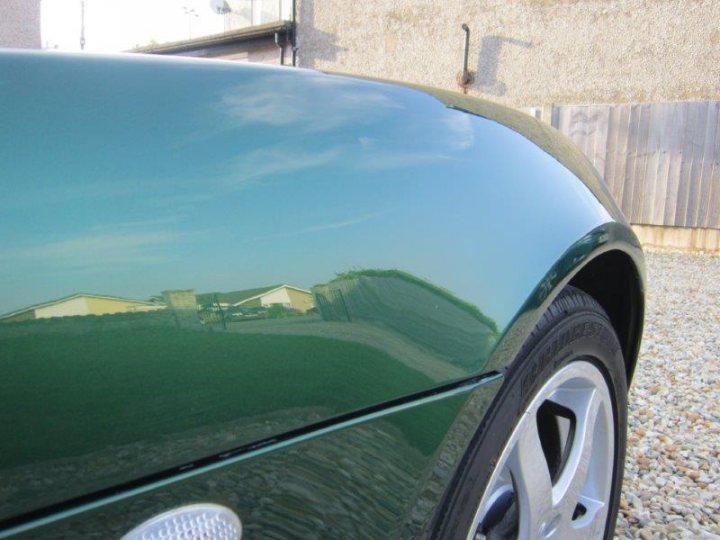 OCD - Page 1 - Elise/Exige/Europa/340R - PistonHeads - The image shows the backside of a car with a glossy green paint finish. The car is parked on a pebble driveway, and the clear sky reflects in the car's surface. There's a building and a white gate with wooden planks visible behind the car. The photo has a shallow depth of field, focusing on the car's rear wheel while the rest of the environment is slightly blurred.