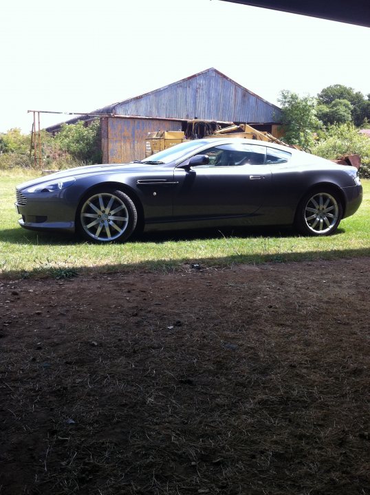 Pistonheads - In the tranquil picture, a sleek, silver Aston Martin sports car commands attention. It's parked on a lush, green lawn, surrounded by the soft scenery of a sunlit garden. The car's position and the stretching shadow suggest it's been parked under a structure, possibly a barn or outdoor shelter. The day outside is pleasant, with the building in the background subtly enhancing the pastoral ambiance of the scene.