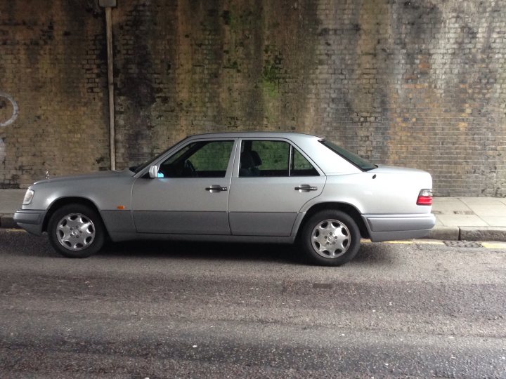 Show us your SIDE! - Page 29 - Readers' Cars - PistonHeads - The image shows a light-gray sedan parked at the side of a road. The car is positioned slightly off the center line, adjacent to a brick wall. The setting appears to be an urban environment, possibly a street with less traffic. There are no people in the scene, giving the photo a quiet, still atmosphere. There are some smaller objects in the picture which could be trash or street furniture, but they don't detract from the car itself, which is the main focus. The car is in good condition, with no noticeable damage or wear.