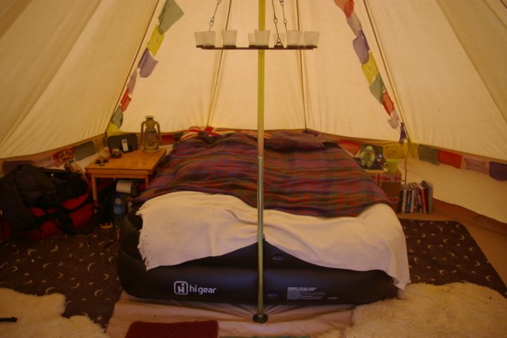 Anyone else got a bell tent ? - Page 1 - Tents, Caravans & Motorhomes - PistonHeads - This image features a cozy, inside-of-a-tent setting. There is a neatly made bed with a red and blue plaid blanket and a white fringed throw blanket. A roof hatch is visible, providing a ceiling for the space. On the floor next to the bed, there is a wardrobe, and opposite the bed is a table with a tent footprint written on it. Additionally, there are small items like cups and books scattered around the area.