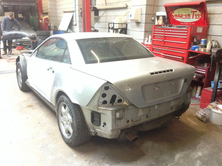 SLK R170 front wings - Page 1 - Mercedes - PistonHeads - This image displays a partially disassembled white compact car parked inside a garage-like space. The car's rear end is missing its bumper, exposing the chrome exhaust pipe. Bare bricks make up the wall in the background, hinting at a workshop environment. Red toolboxes and utensils are visible, reinforcing the impression of a garage or repair workshop. The overall scene suggests a repair or maintenance process is underway.